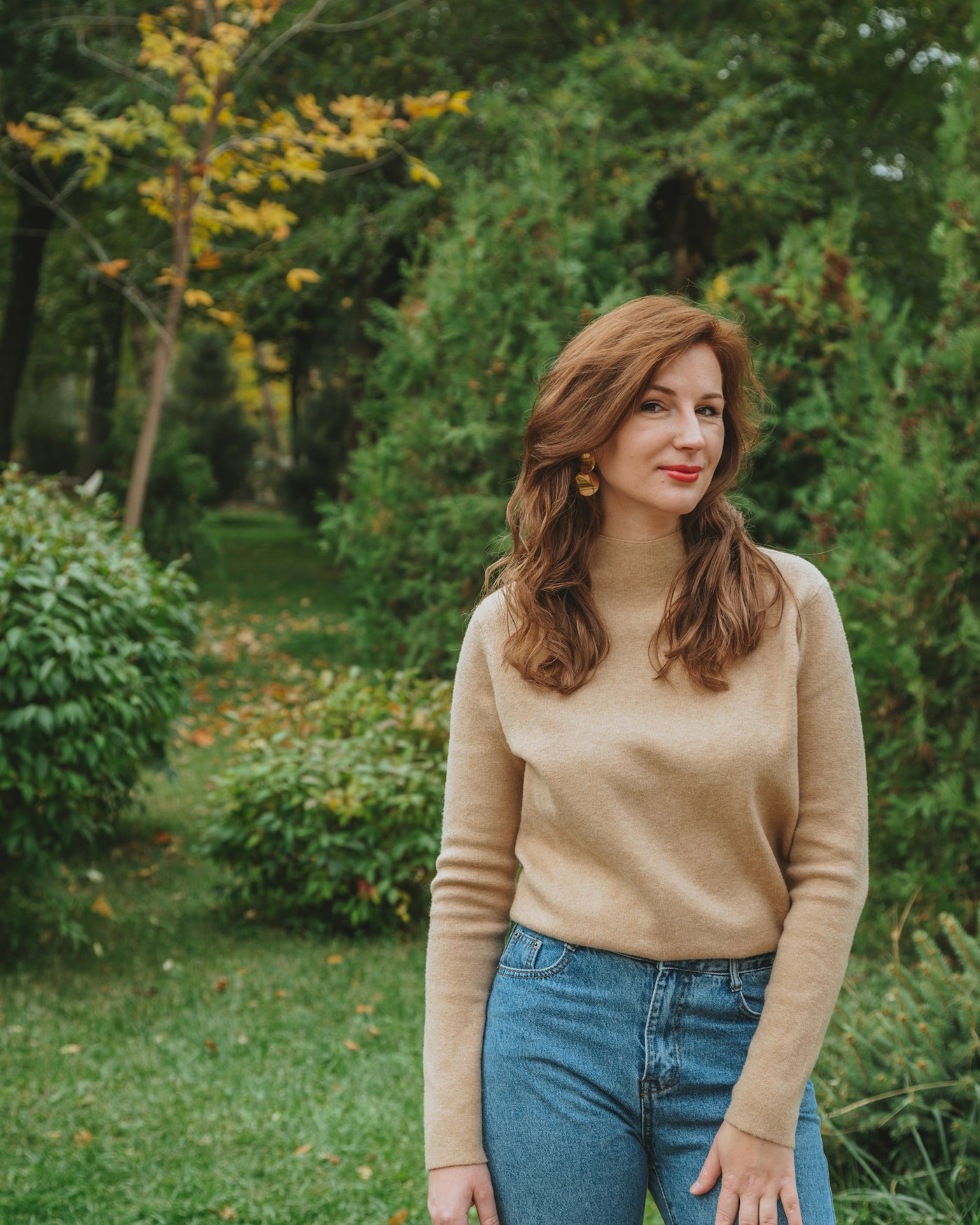 Camel crewneck sweater with medium wash straight leg jeans and gold drop earrings, standing in a garden in warm daylight