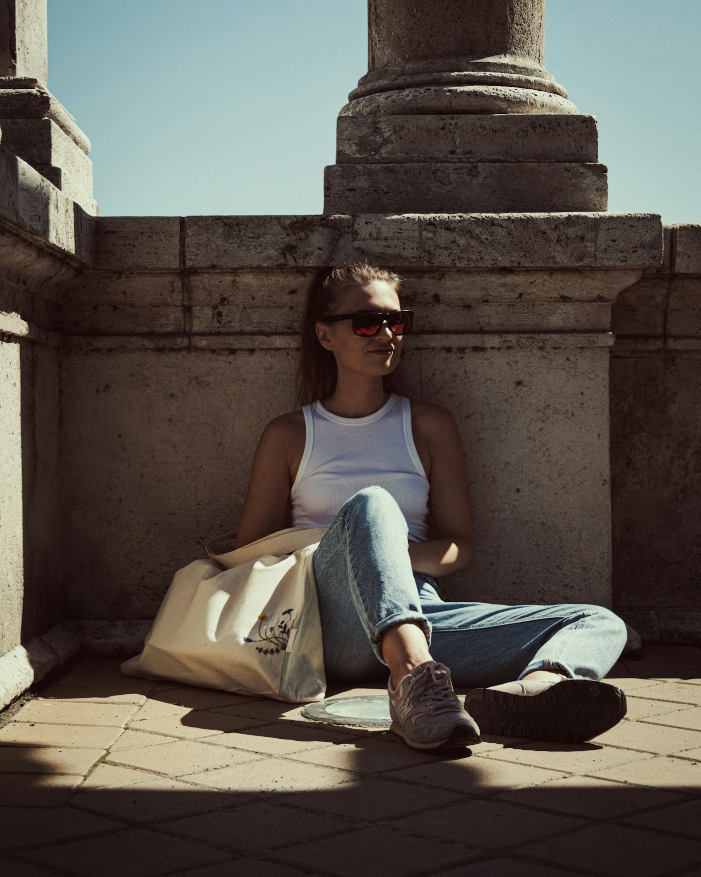 White ribbed tank with wide leg light denim jeans and a cream canvas tote, resting in the shade of stone columns