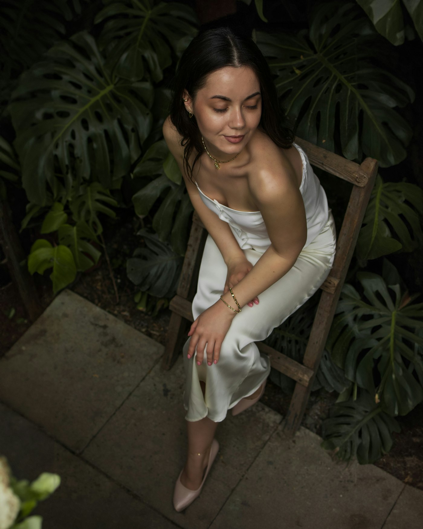 Ivory satin slip midi dress with a fine pendant necklace and a small gold earring, seated in a lush garden in soft natural light